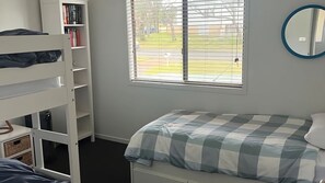 Quarto com beliche, mesa de cabeceira, estante de livros, janela com persianas e espelho redondo na parede.