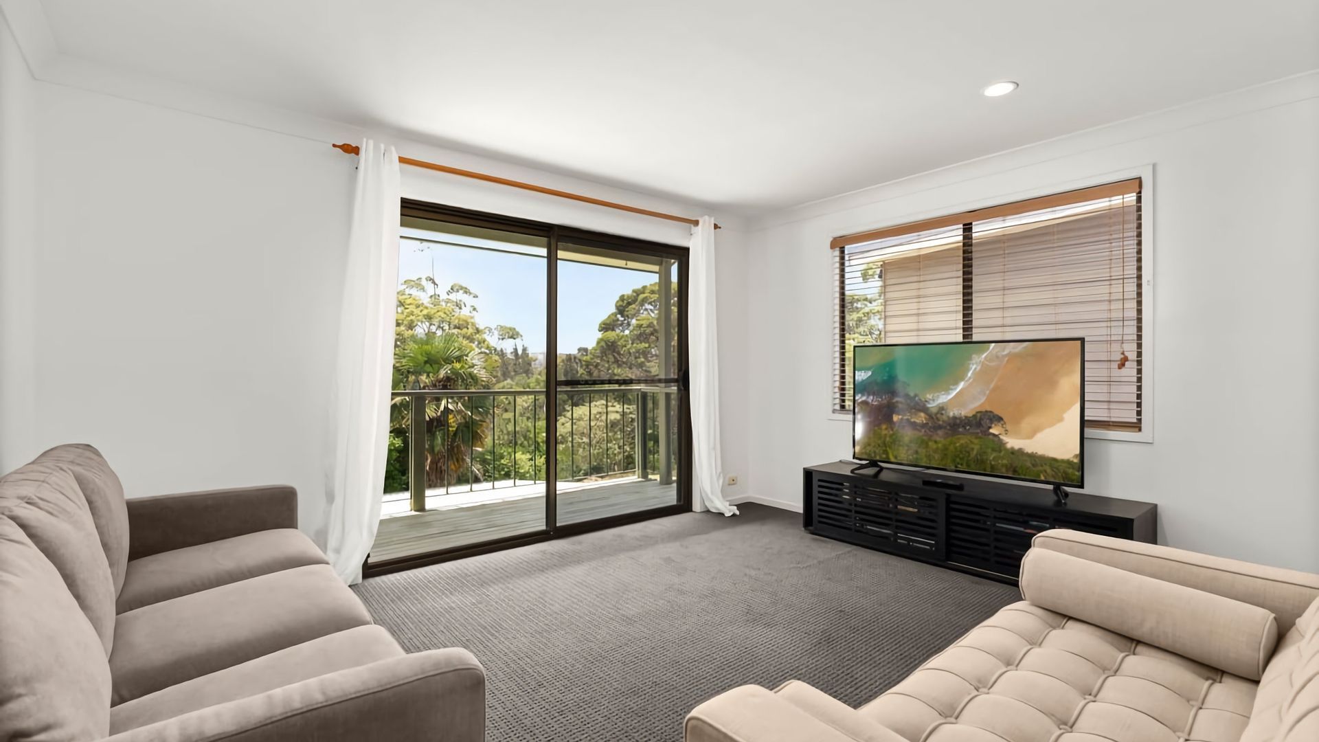 A living room with a large TV, a sofa, and a balcony with a view of trees.