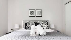 A neatly made bed with a white quilt, two patterned pillows, and rolled white towels on the bed.