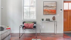 A room with a wooden floor, a grey sofa, a standing chair with a pink cushion, a small white console table with a plant, and a framed painting above the console table.