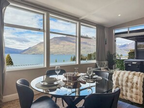 A dining area with a table set for four, large windows with a mountain and lake view, and a cozy seating area with a fur throw. - Chic Central Queenstown Retreat- Newly Renovated (Queenstown)