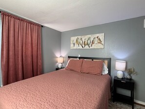 A bedroom with a large bed, two bedside tables with lamps, a wall-mounted artwork of birds, and a window with a curtain.