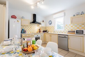 A kitchen with a checkered yellow and white backsplash, a black range hood, and a dining table set with fruits and dishes. - Maison 3 chambres Lac, Jardin  et  terrasse (BISCARROSSE)