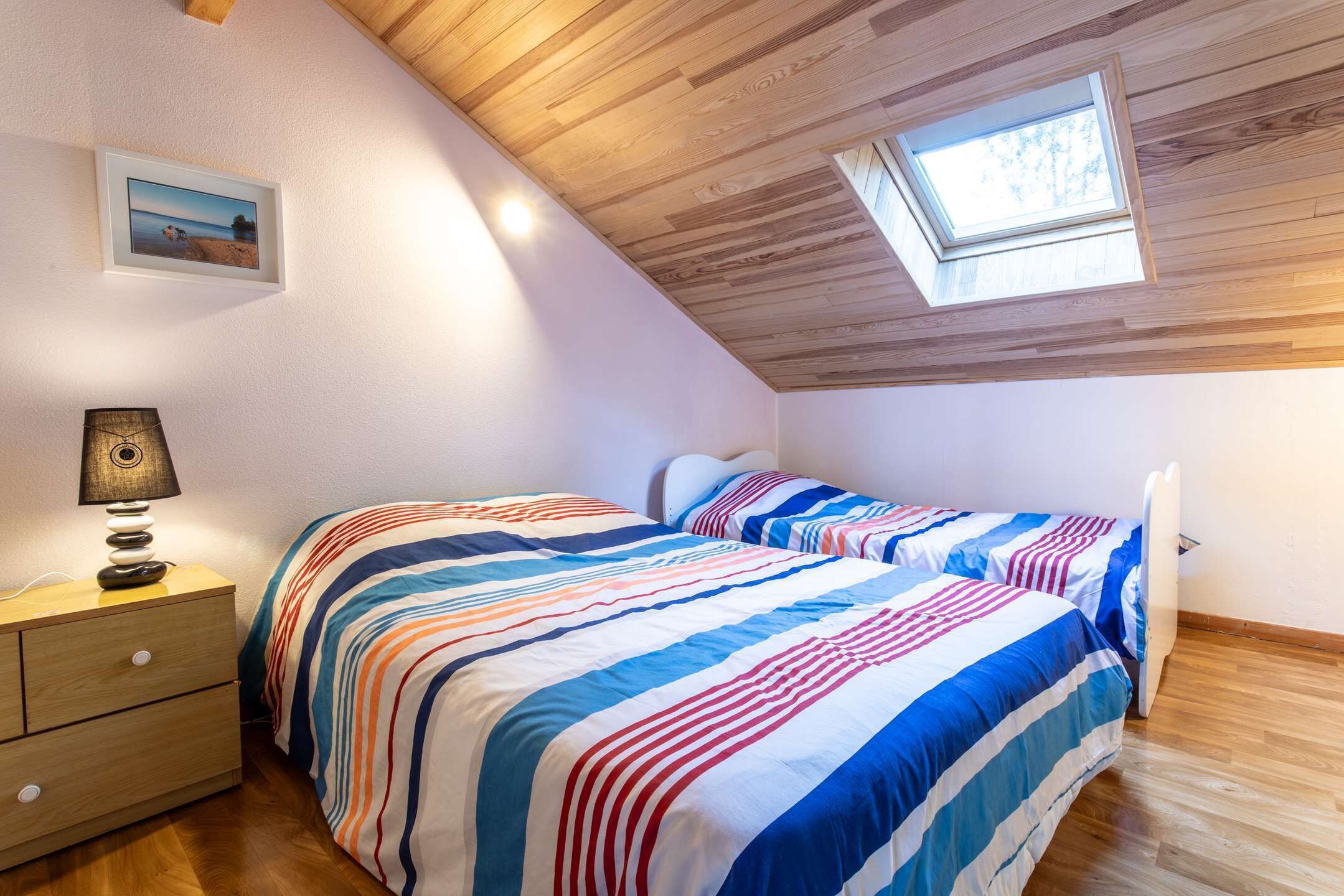 Camera da letto con due letti, soffitto in legno, un lucernario, una lampada da comodino e un quadro appeso al muro.