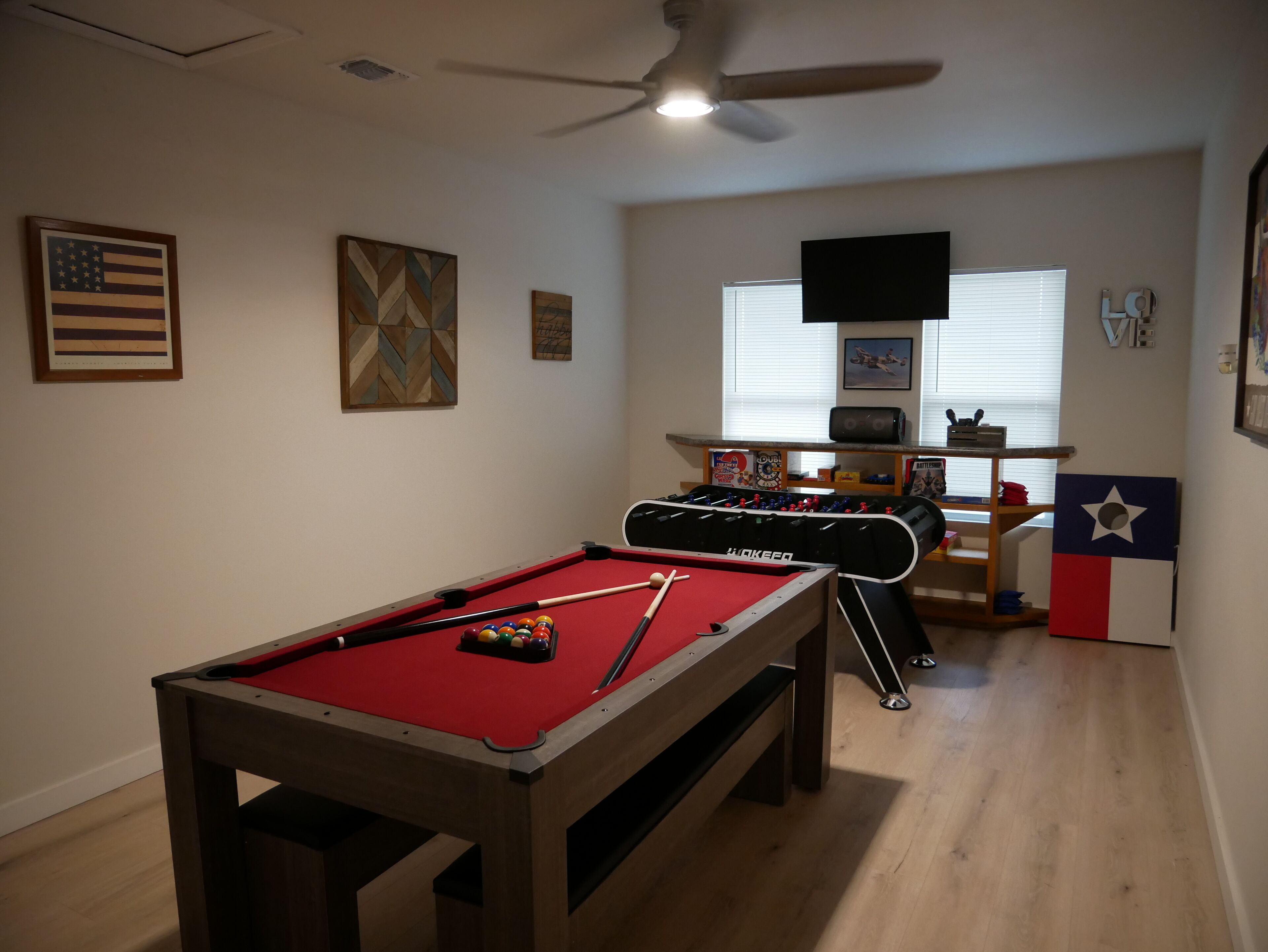 Een biljart met rode viltkleed, een plafondventilator en een tafelvoetbaltafel in een kamer met houten vloeren en een raam met zonwering.