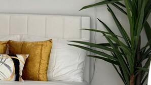A bed with white bedding and gold and white pillows, a wall-mounted light fixture, and a large green plant. - Elegant Hideaway with Free Parking (White Plains)