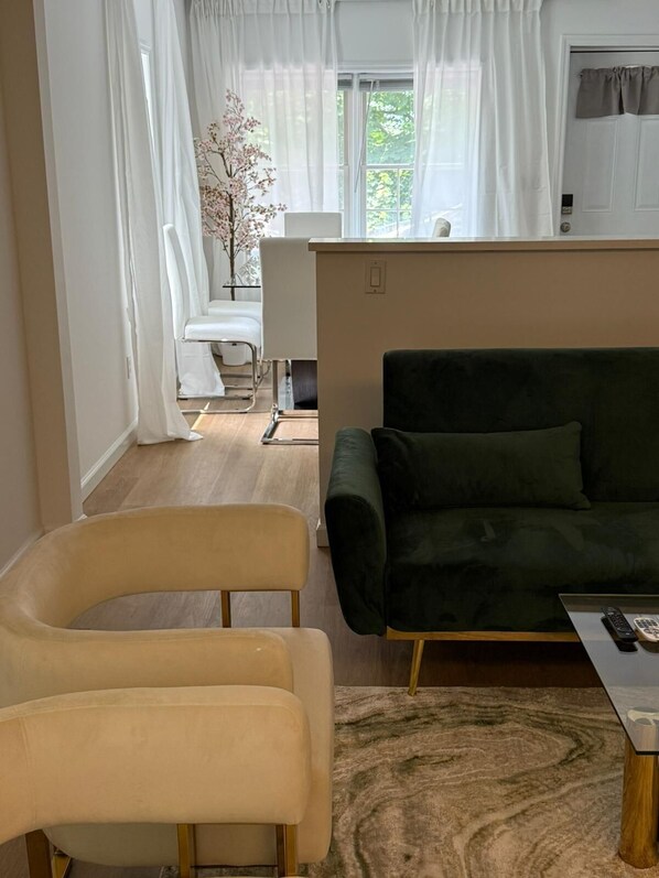 Una sala moderna con un sofá verde, dos sillones beiges, una mesita con un control remoto y una ventana con cortinas traslúcidas.