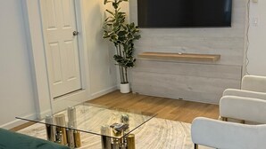 A modern living room with a green sofa, a glass coffee table, a white armchair, and a large flat-screen TV mounted on a light-colored wall. - Elegant Hideaway with Free Parking (White Plains)