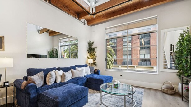 Una sala de estar moderna con un sofá seccional azul, una mesa de centro de vidrio y ventanas amplias que ofrecen vistas a un edificio de ladrillo.