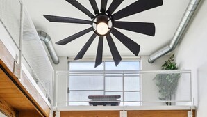 Amplia sala de estar con ventilador de techo alto, ventanas grandes, un sofá azul, una mesa de centro de vidrio y vistas a un edificio.