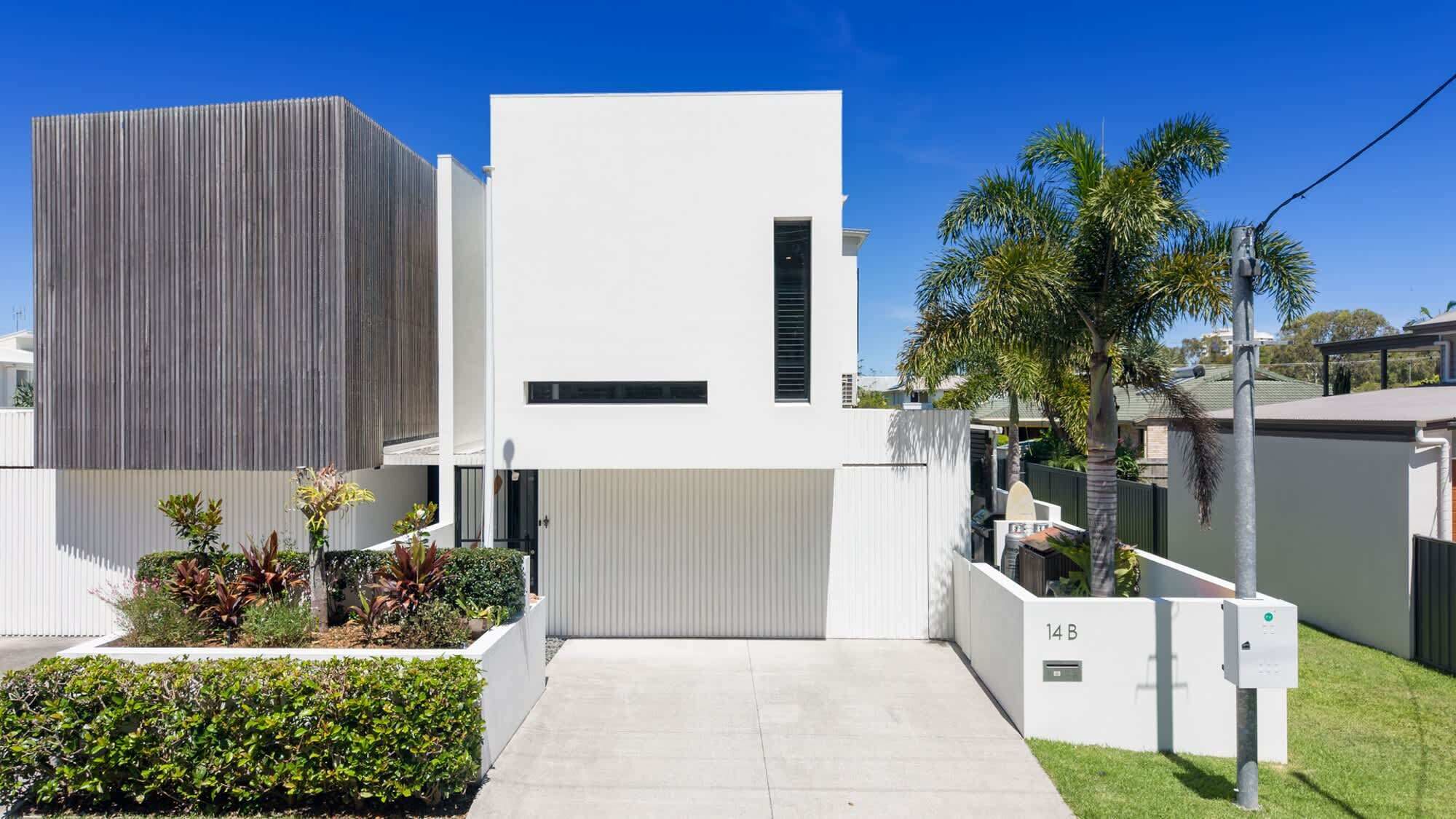 A modern two-story house with a white facade, a large garage door, and a well-maintained garden with various plants.