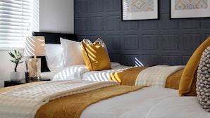Ein ordentlich bezogenes Bett mit weißer und senffarbener Bettwäsche, ein Nachttisch mit Lampe sowie gerahmte Bilder an der Wand.