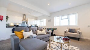 Ein modernes Wohnzimmer mit einem grauen Sofa, einem runden Couchtisch und einem beigefarbenen Sofa. Im Hintergrund ist eine Küche mit weißen Schränken und einer Edelstahl-Dunstabzugshaube zu sehen.