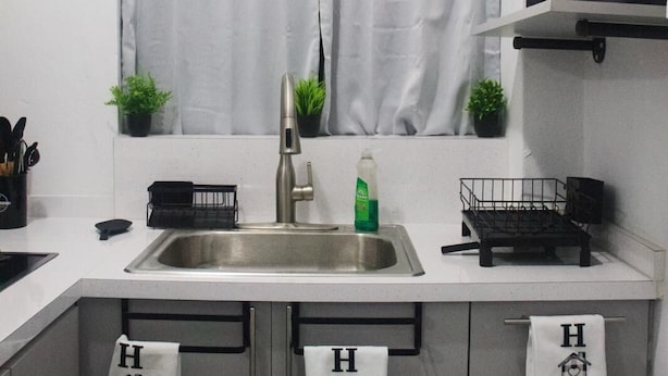 A kitchen with a stainless steel sink, two potted plants, a window with white curtains, and three towels hanging on the cabinet doors. - Unit 1 Cozy Apartment with walking distance of bars, restaurant and beach (san juan)