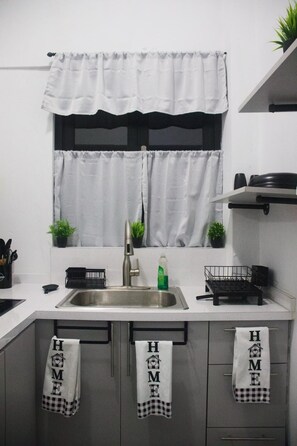 A kitchen with a stainless steel sink, two potted plants, a window with white curtains, and three towels hanging on the cabinet doors.