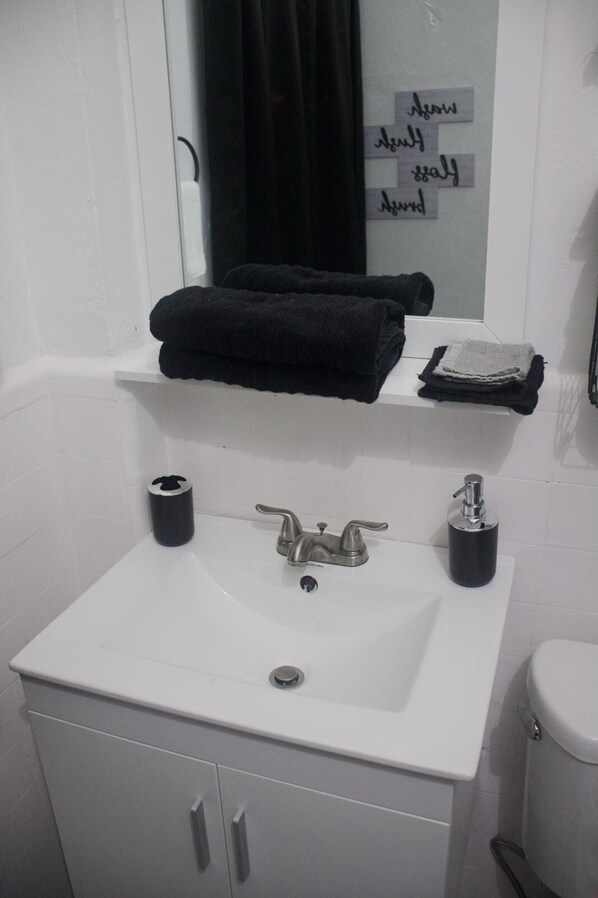A bathroom with a white sink, two black soap dispensers, a mirror with a black towel hanging, and a toilet. - Unit 1 Cozy Apartment with walking distance of bars, restaurant and beach (san juan)