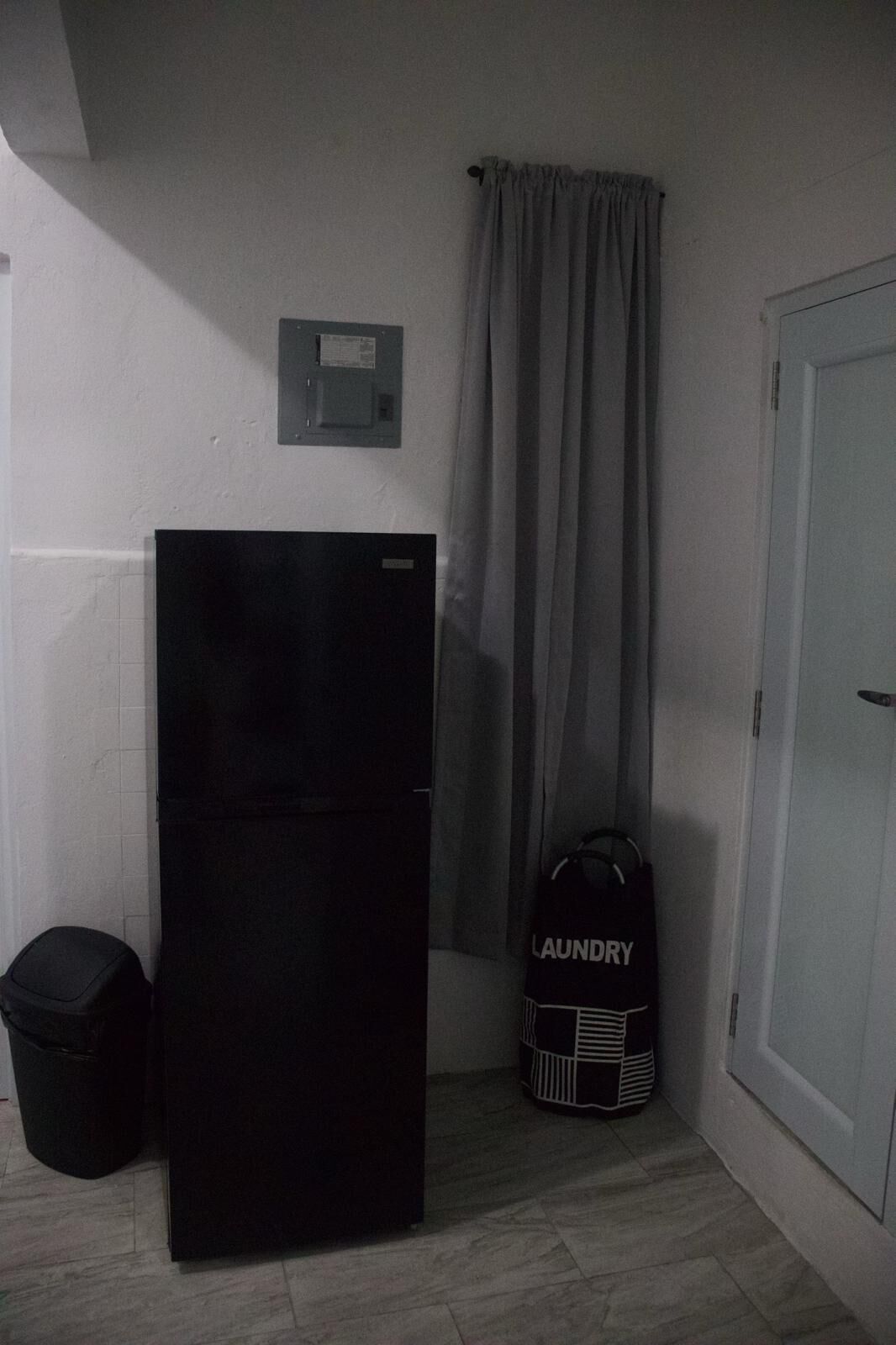 A compact room with a black refrigerator, a trash bin, a laundry bag, and a door.