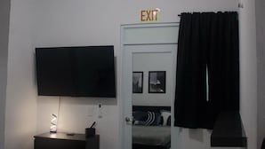 A small, dimly lit room with a bed, a TV, a door with an exit sign, and a small trash bin. - Unit 1 Cozy Apartment with walking distance of bars, restaurant and beach (san juan)