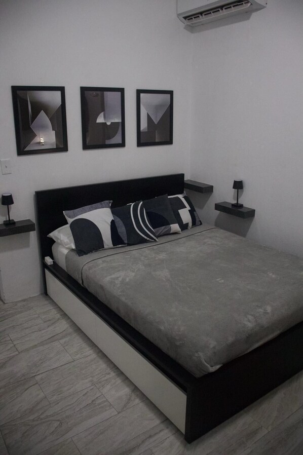 A modern bedroom with a black headboard, grey bedding, and three framed artworks on the wall.