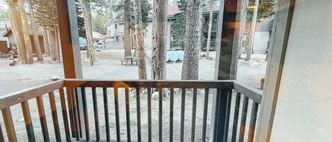 A wooden deck with a metal railing, overlooking a parking area with trees and buildings in the background.