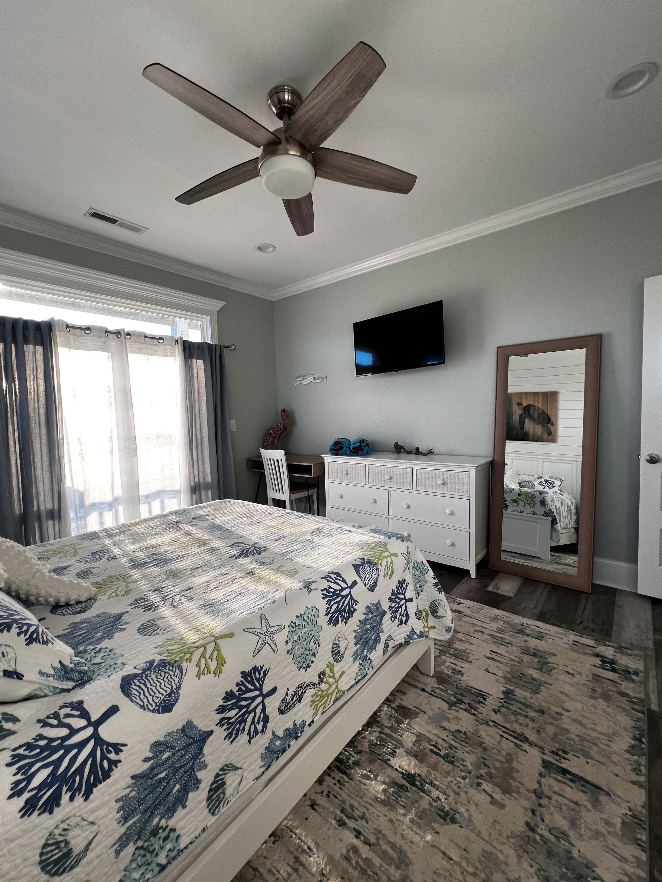Ein Schlafzimmer mit Bett, Schreibtisch, Kommode, Fernseher und Deckenventilator.