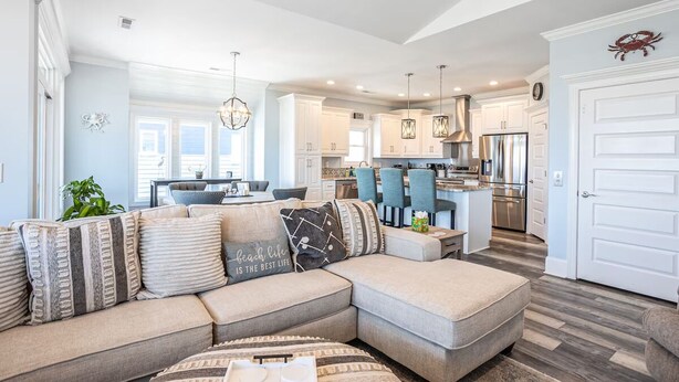 A modern living room with a sectional sofa, a dining area, and a kitchen with white cabinetry and stainless steel appliances. - Top Sail Beach Sunsets (Sneads Ferry)