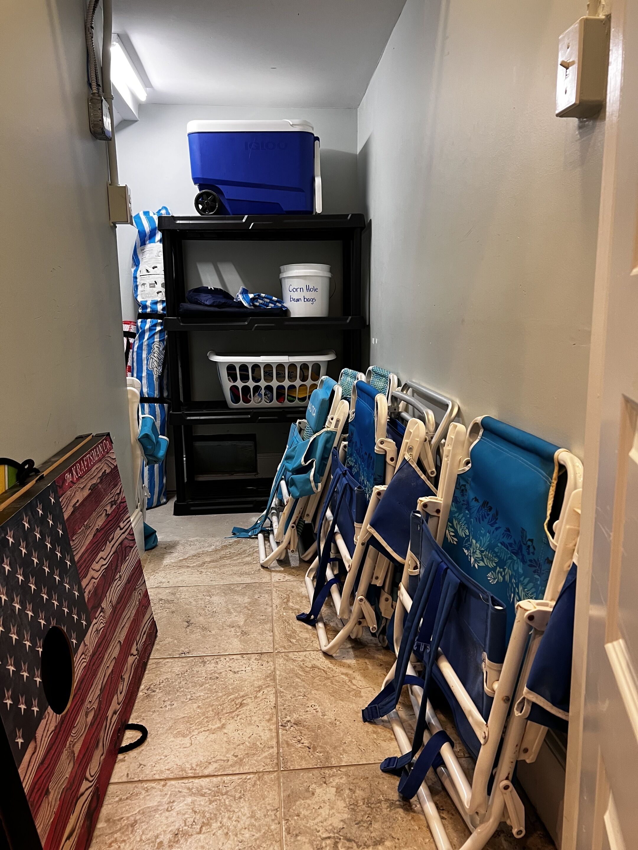 A narrow hallway with a row of blue and white baby cribs lined up against the wall, a laundry basket, and a shelf with a container labeled "Corn Hole bean bags".