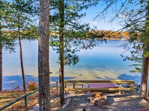Eine Holzterrasse am Seeufer mit Feuerstelle und Bänken, umgeben von Bäumen und mit Blick auf einen ruhigen See.