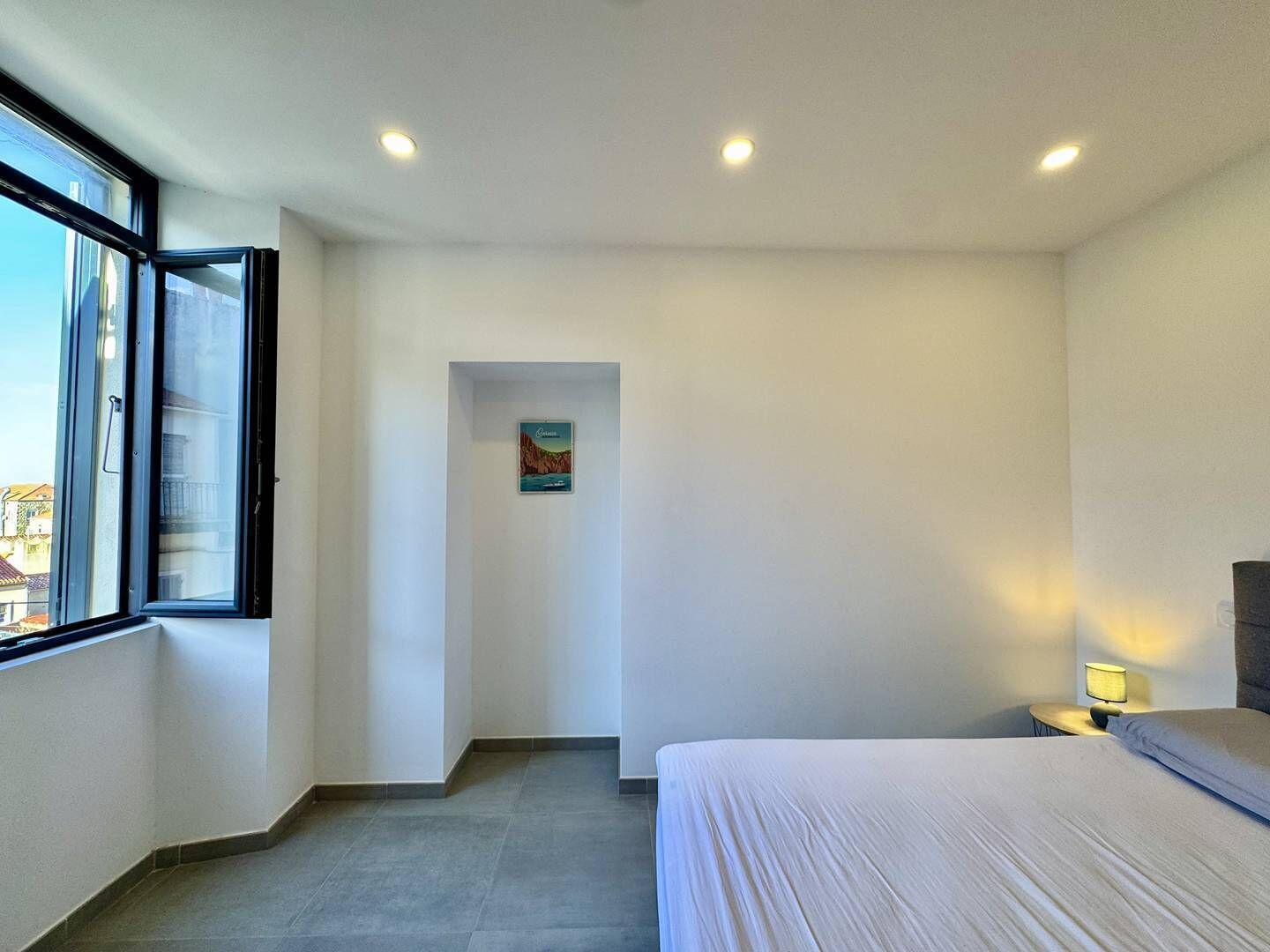 Ein Schlafzimmer mit einem großen Bett, einem Nachttisch mit Lampe, einem Fenster mit Jalousien und einer Tür zu einem anderen Raum.