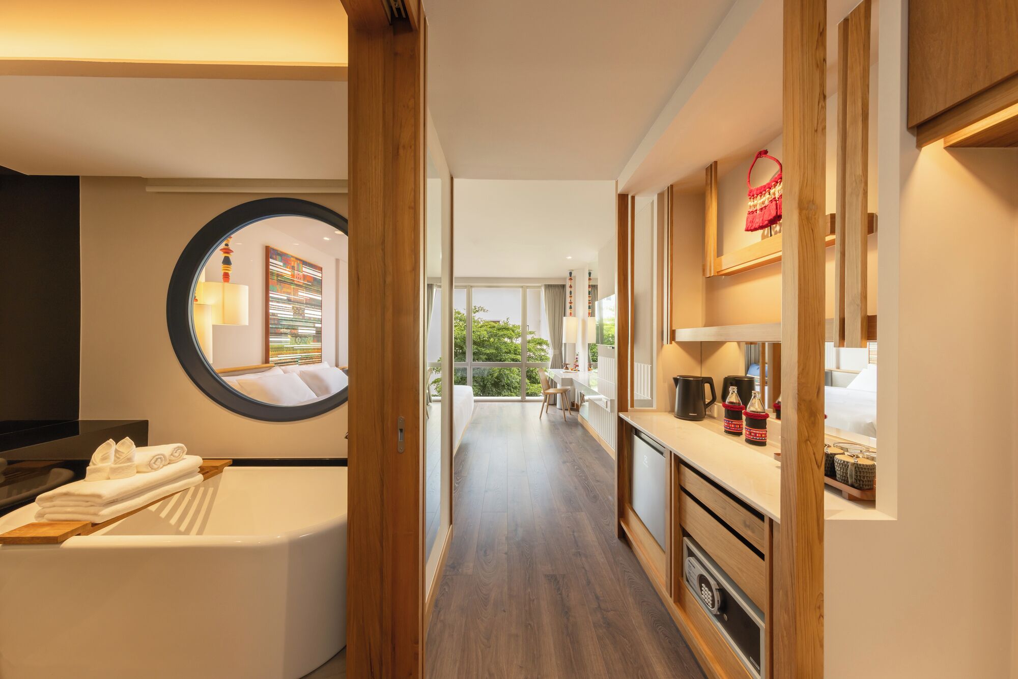 A hotel room with a circular mirror, a bathtub, a wooden cabinet, and a view of the outdoors.