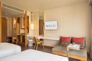 A hotel room with a bed, a desk, a chair, a sofa with cushions, a trash bin, and a wall-mounted TV displaying 'NAU'. - Na U Hotel (Chiang Mai)