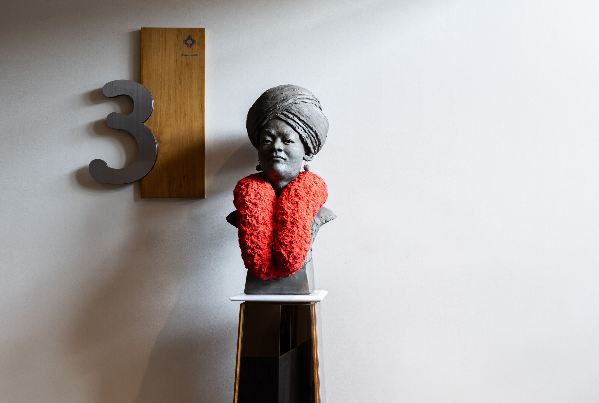 Uma escultura com um objeto vermelho texturizado sobre um pedestal, um painel de madeira com um número e uma parede lisa.