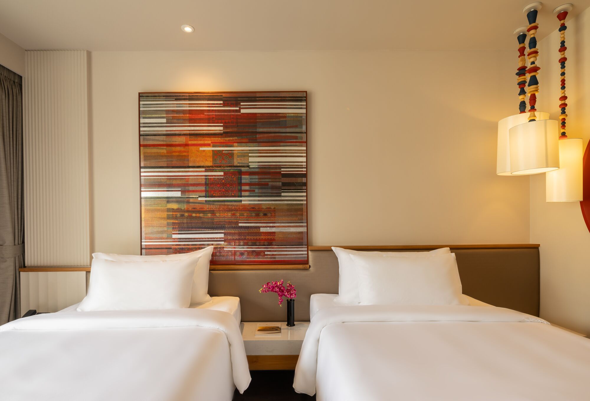 A hotel room with two beds, a large abstract painting, a vase with flowers, and a wall-mounted lamp.