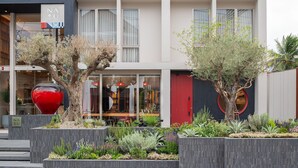 Un edificio moderno con una puerta roja, un gran jarrón rojo y diversas plantas en primer plano.
