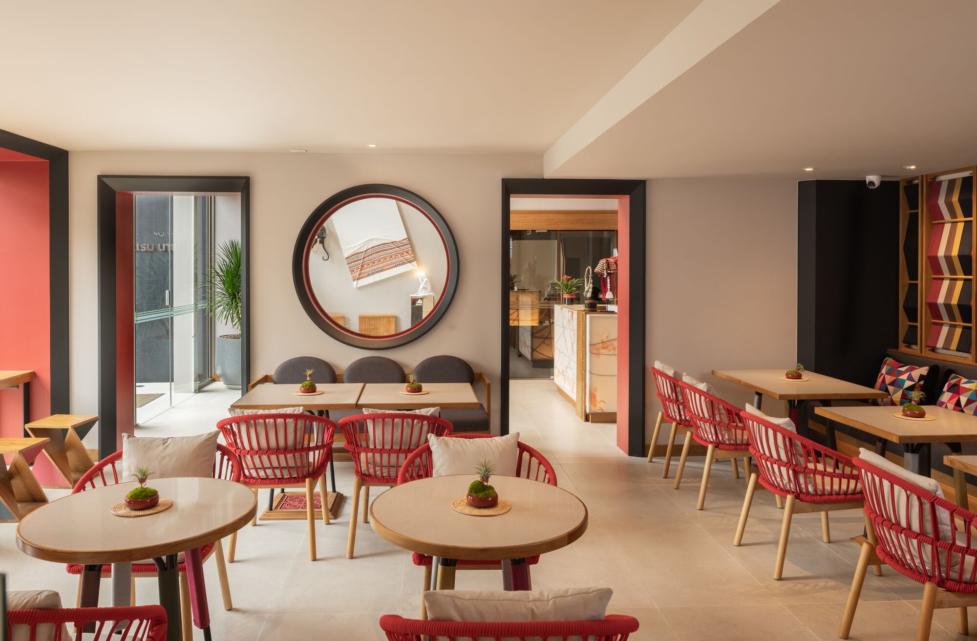 A modern dining area with round tables and red wicker chairs, a large mirror on the wall, and a view into the kitchen area.