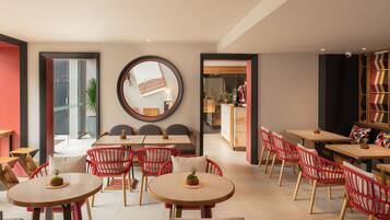 A modern dining area with round tables and red wicker chairs, a large mirror on the wall, and a view into the kitchen area.