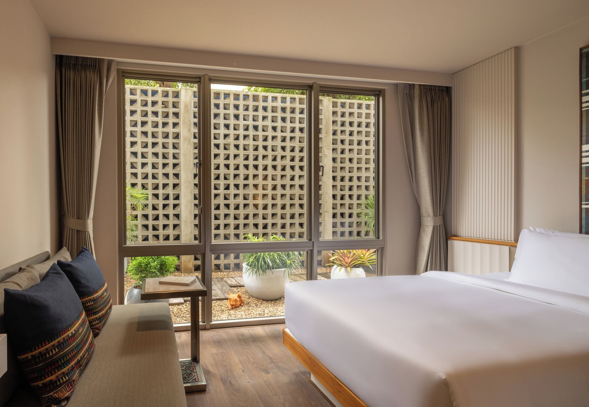 Habitación de hotel con una cama grande, una zona de estar con cojines, una mesita y vistas a trav és de una ventana con celosía.