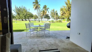 Un patio abierto con una mesa y sillas, vista a un área verde con palmeras y un coche estacionado cerca.