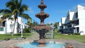 A multi-tiered fountain with water cascading down, surrounded by a tiled pool, palm trees, and modern buildings. - Playas del Yunque - Beach Front - Rio Grande, PR (Rio Grande)