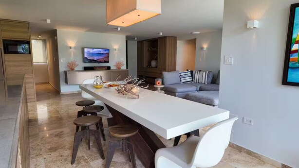 A modern kitchen with a central island, bar stools, a grey sofa, and a TV mounted on the wall. - Playas del Yunque - Beach Front - Rio Grande, PR (Rio Grande)