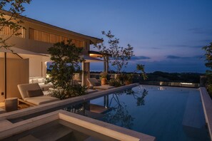 Ein moderner Poolbereich im Freien mit Liegestuhl und Sofa, umgeben von Pflanzen und Blick auf die Stadt im Abendlicht.