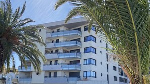 Un immeuble résidentiel à plusieurs étages, doté de balcons, avec des voitures garées et des palmiers.