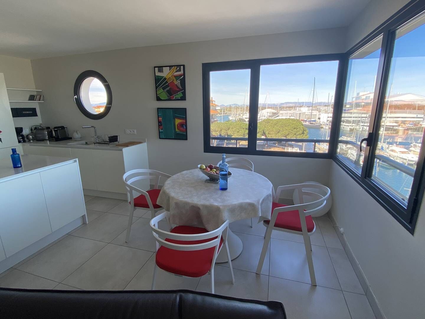 Une cuisine moderne avec des meubles blancs, une fenêtre ronde et un coin repas comprenant une table et des chaises.