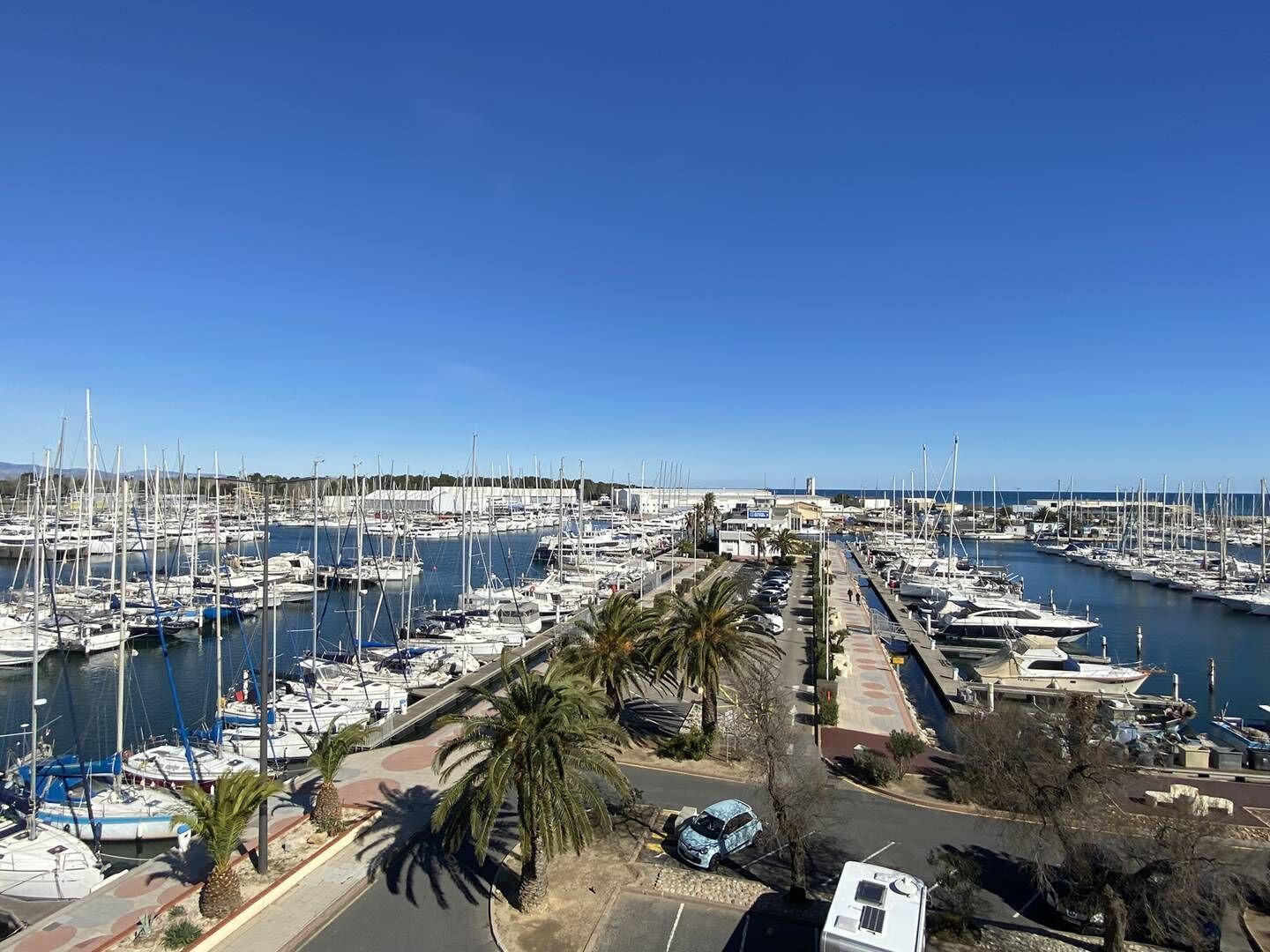 Un puerto deportivo con numerosos barcos amarrados, una zona de estacionamiento y un cielo azul despejado.