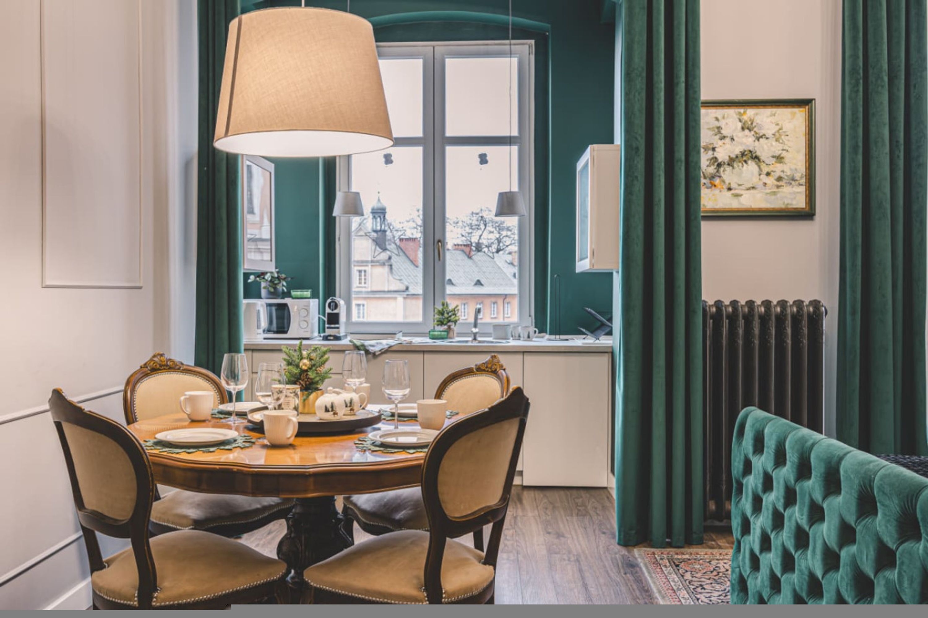 A dining area with a round table, chairs, a window with a view of buildings, a radiator, and a green upholstered bench.