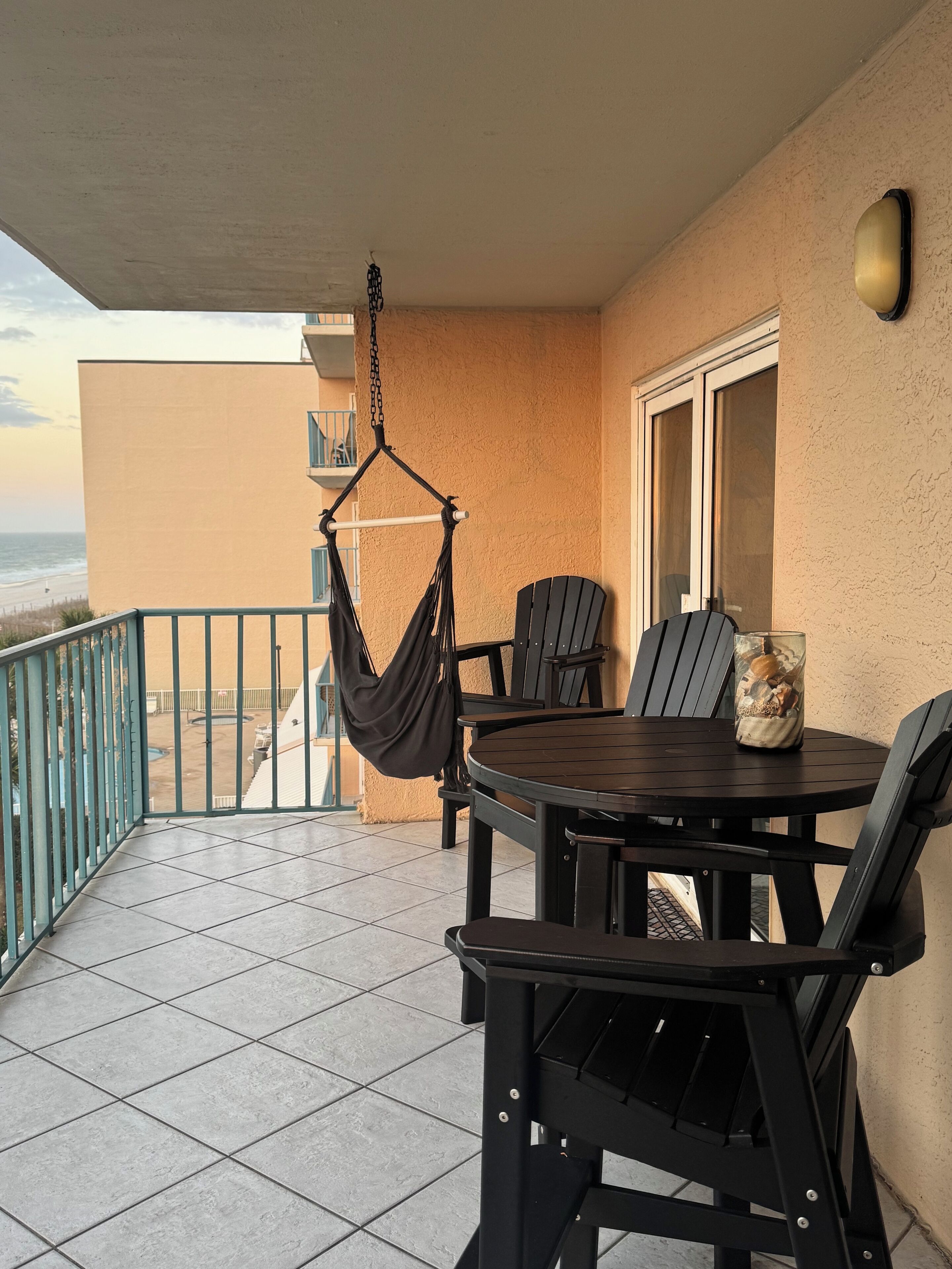 A balcony with a hanging chair, a round table with chairs, and a view of the beach.