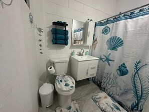 A bathroom with a toilet, sink, and mirror. There is a towel rack with a blue towel, a trash bin, and a shower curtain with sea creature designs. - Unit 3 Cozy Apartment with walking distance from bars, restaurants and beach (San Juan)