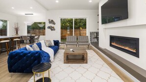 A modern living room with a blue sofa, grey chairs, a wooden coffee table, and a fireplace. - Brand New 5BR Home Fenced Yard Ample Parking (Renton)