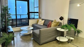 Een moderne woonkamer met een grijze bank, een klein rond tafeltje, een groot raam met uitzicht op gebouwen en verschillende potplanten.