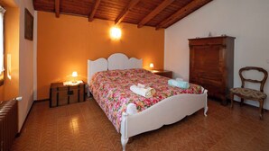 Ein Schlafzimmer mit Holzbalkendecke, einem Bett mit Blumenmuster-Bettwäsche, einem Nachttisch, einem Holzschrank und einem Sessel.